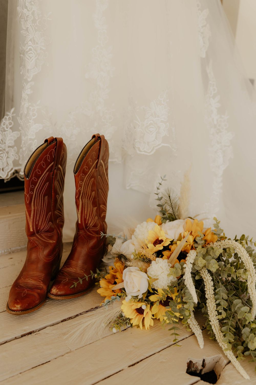 Brown leather cowboy boots sit beside yellow sunflowers and white flowers on a wooden floor.