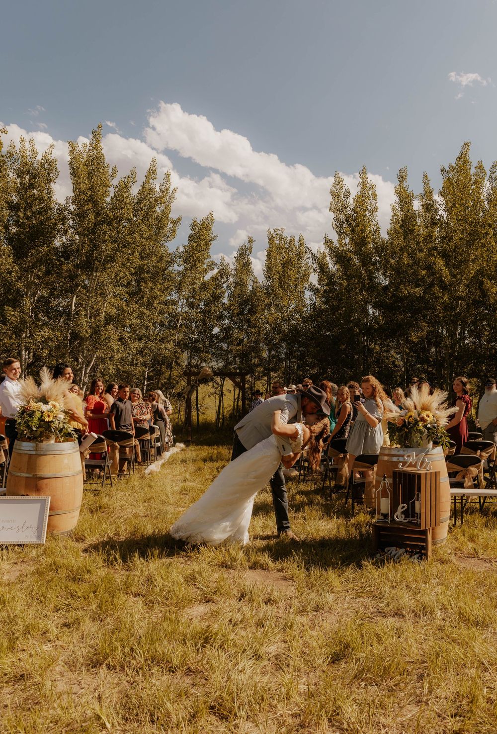 Sunny outdoor wedding ceremony with guests seated in chairs among trees.