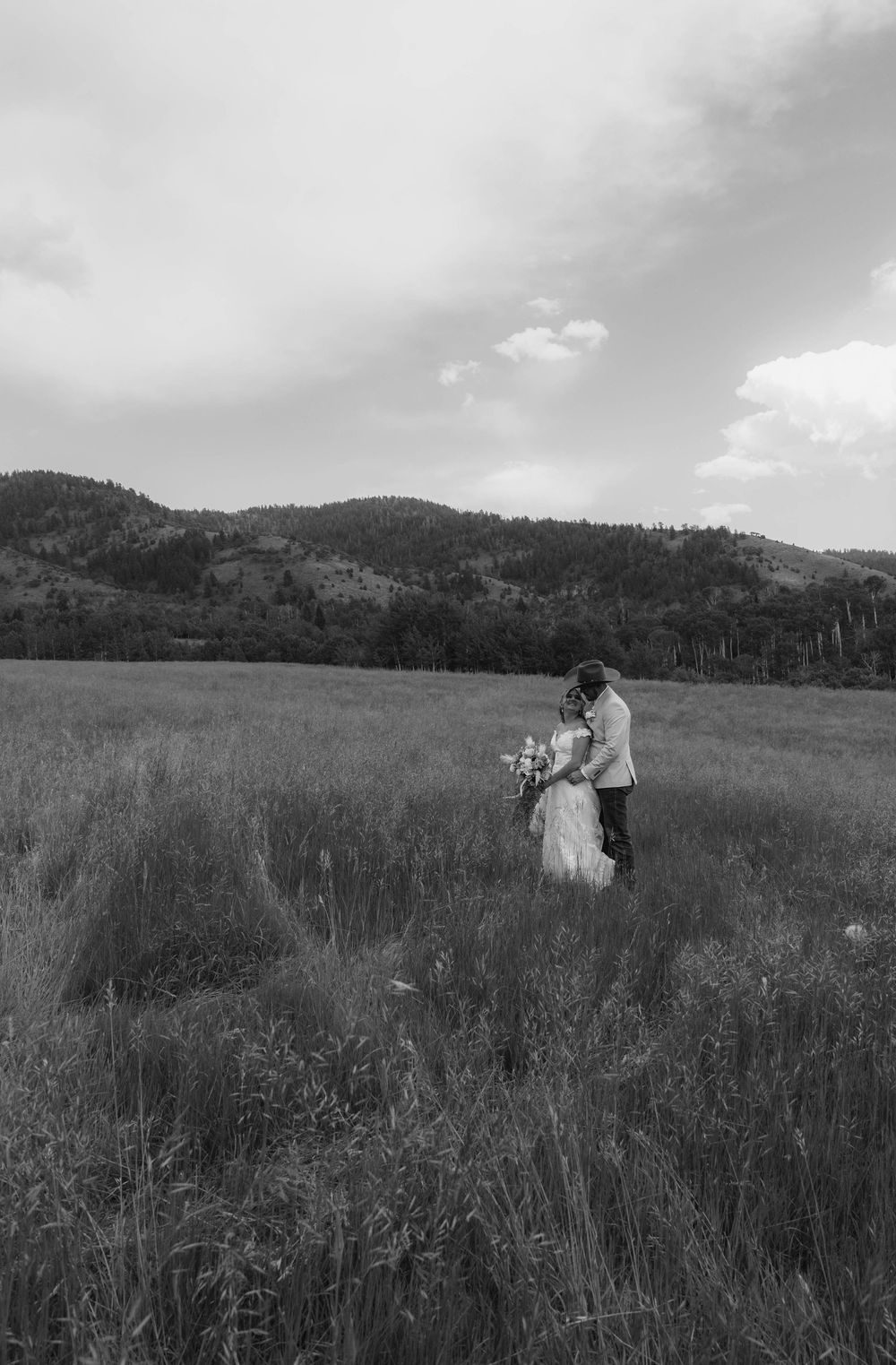 Black and white photo of a western couple embracing in a meadow with mountains in background.