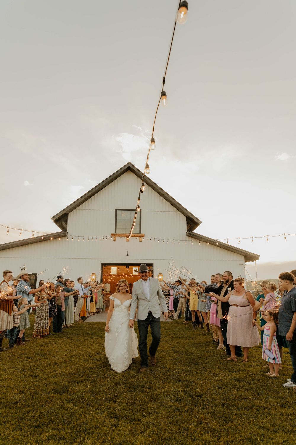 Outdoor western wedding celebration at dusk with string lights illuminating a white barn venue.