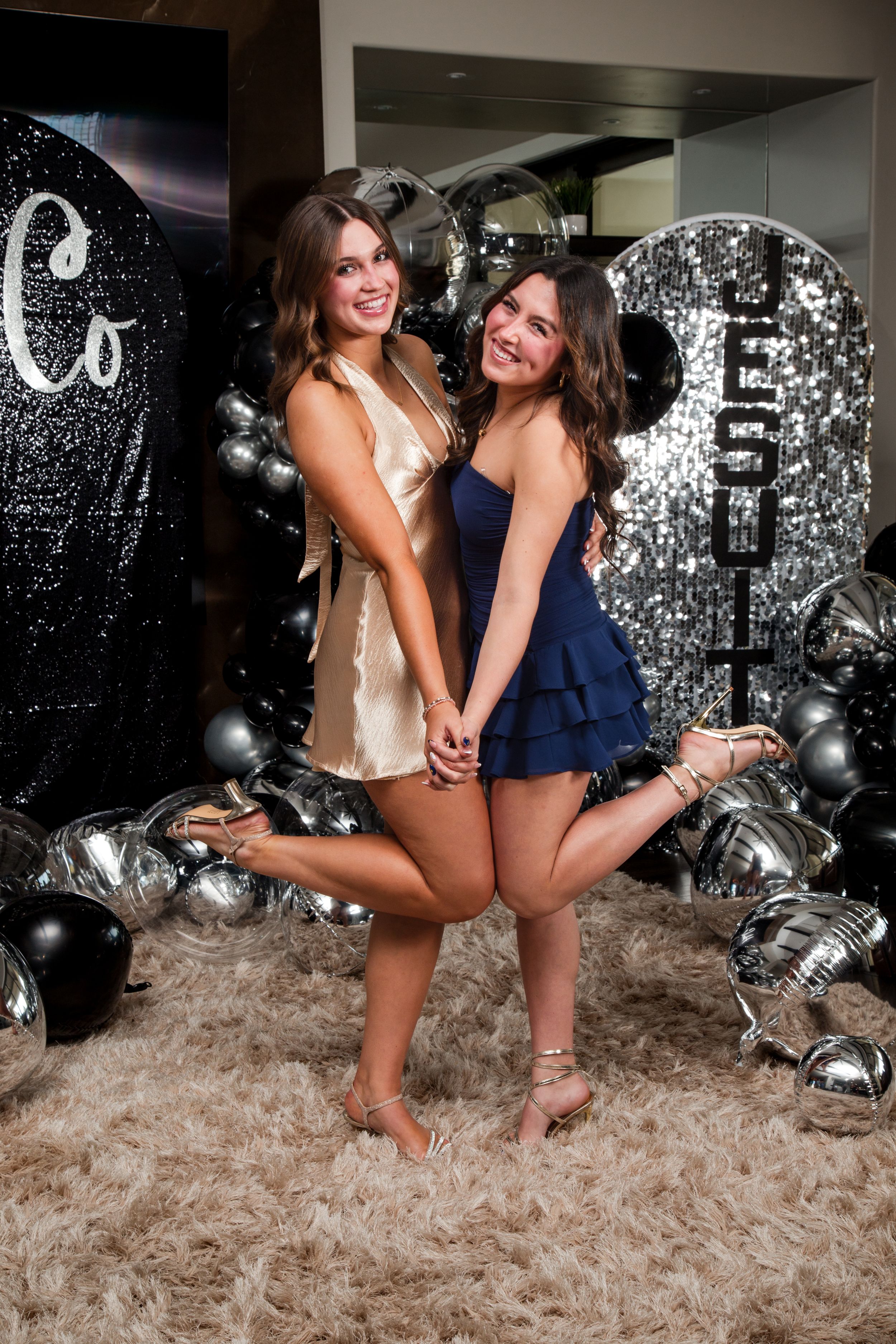 Two friends in party dresses pose together on a fluffy rug with silver balloons in the background. homecoming sacramento california