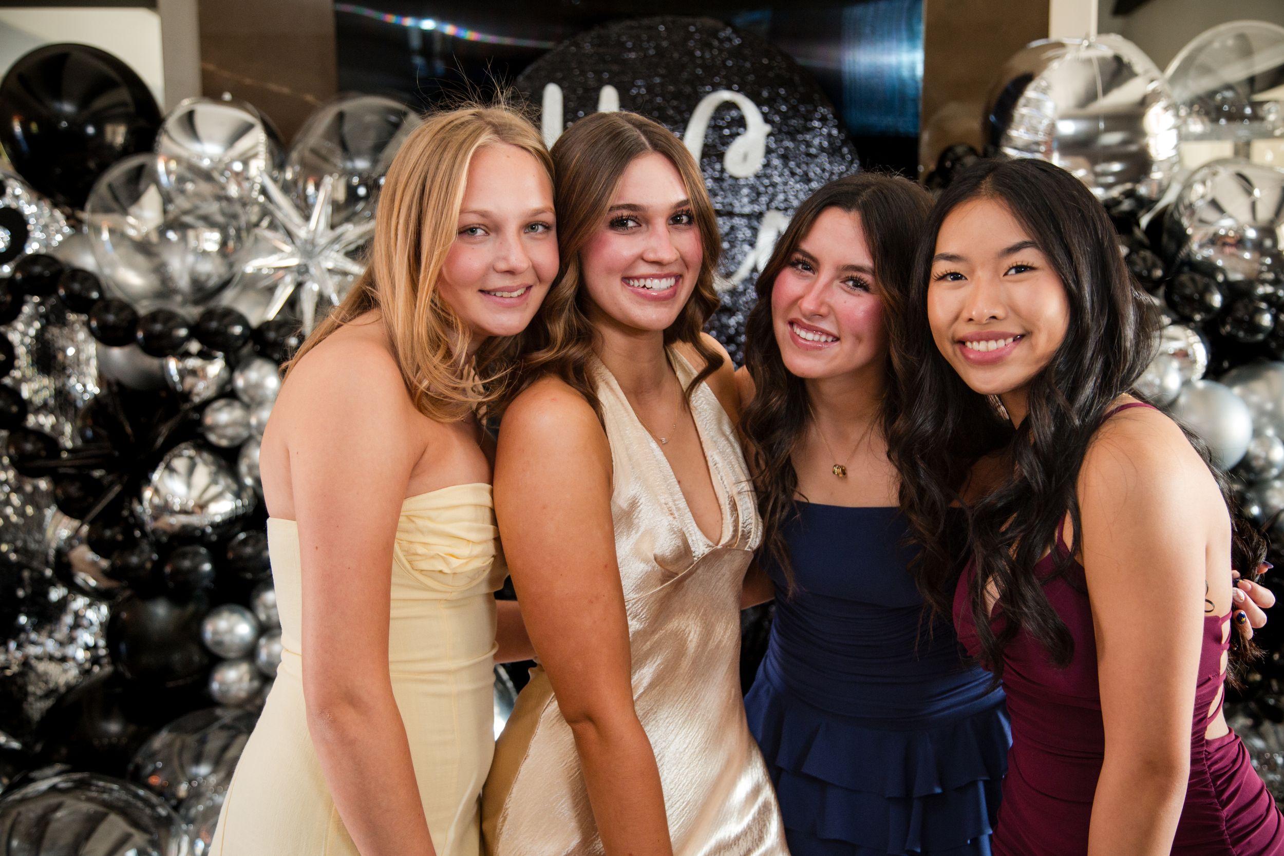 A group of girls poses together at a formal event wearing elegant dresses in various colors against a sparkling backdrop. homecoming