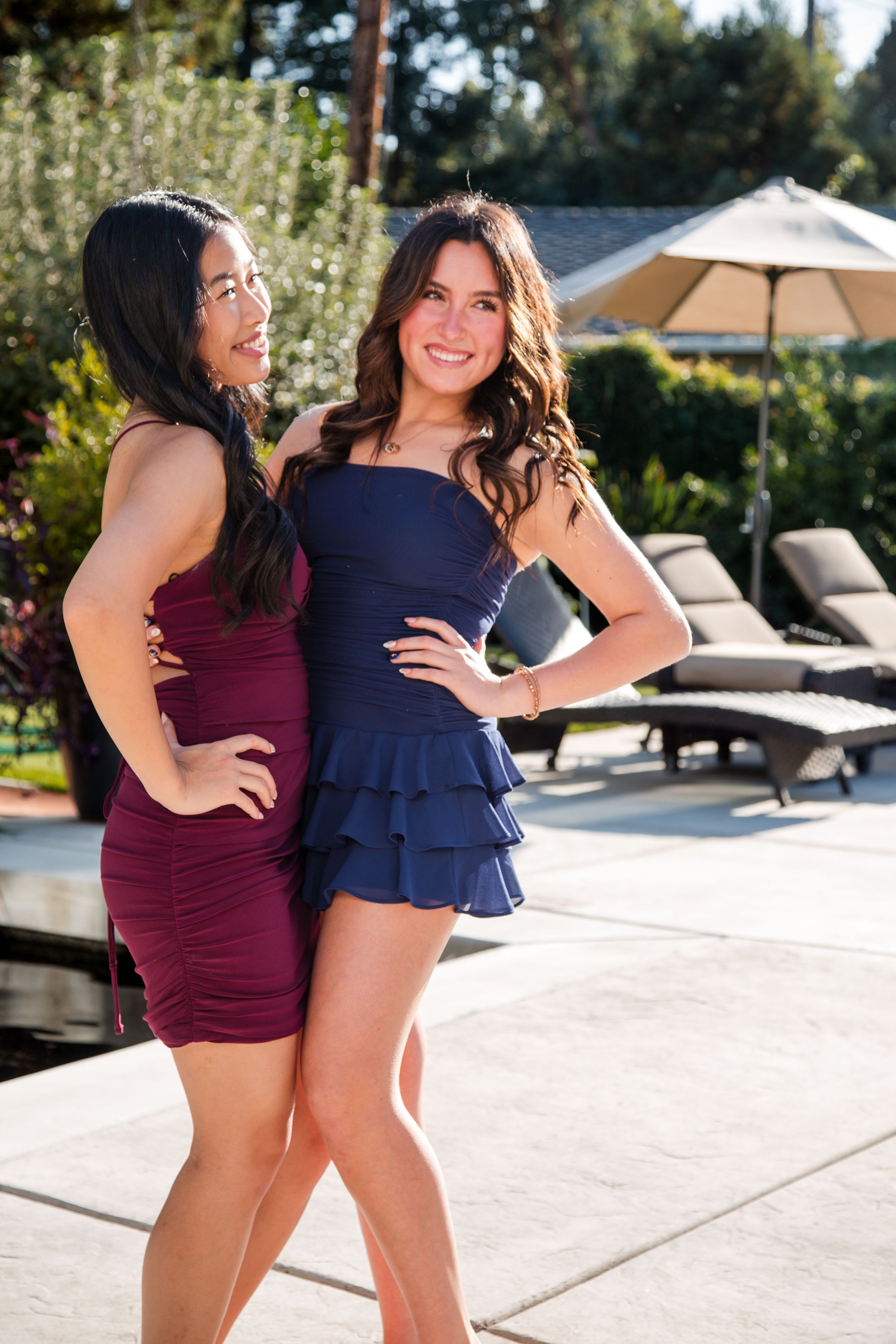 Two friends in navy and burgundy dresses pose playfully by a pool on a sunny day. homecoming pics sacramento photographer