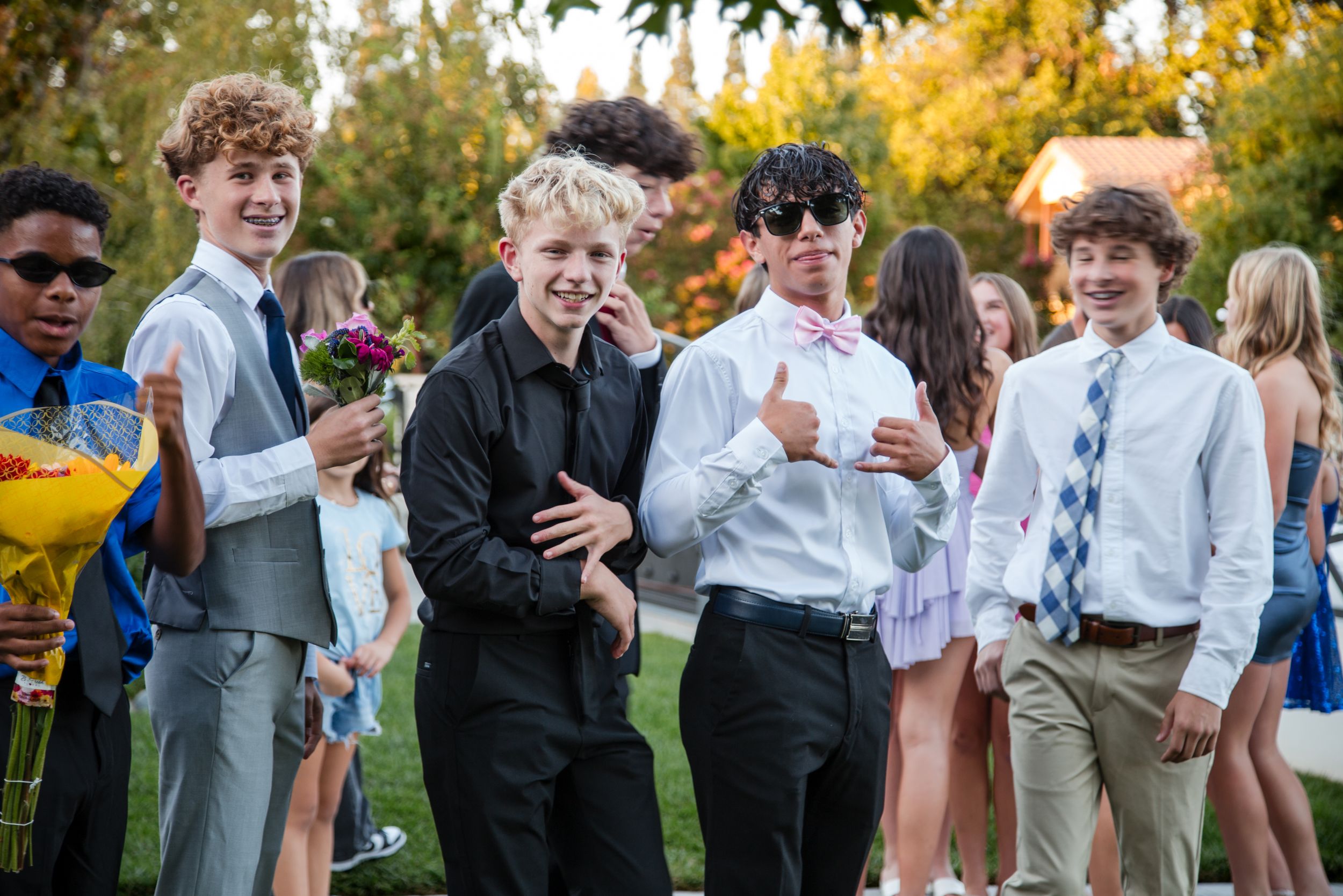 A group of young people in formal attire gather outdoors during an event with autumn foliage in the background. homecoming photos