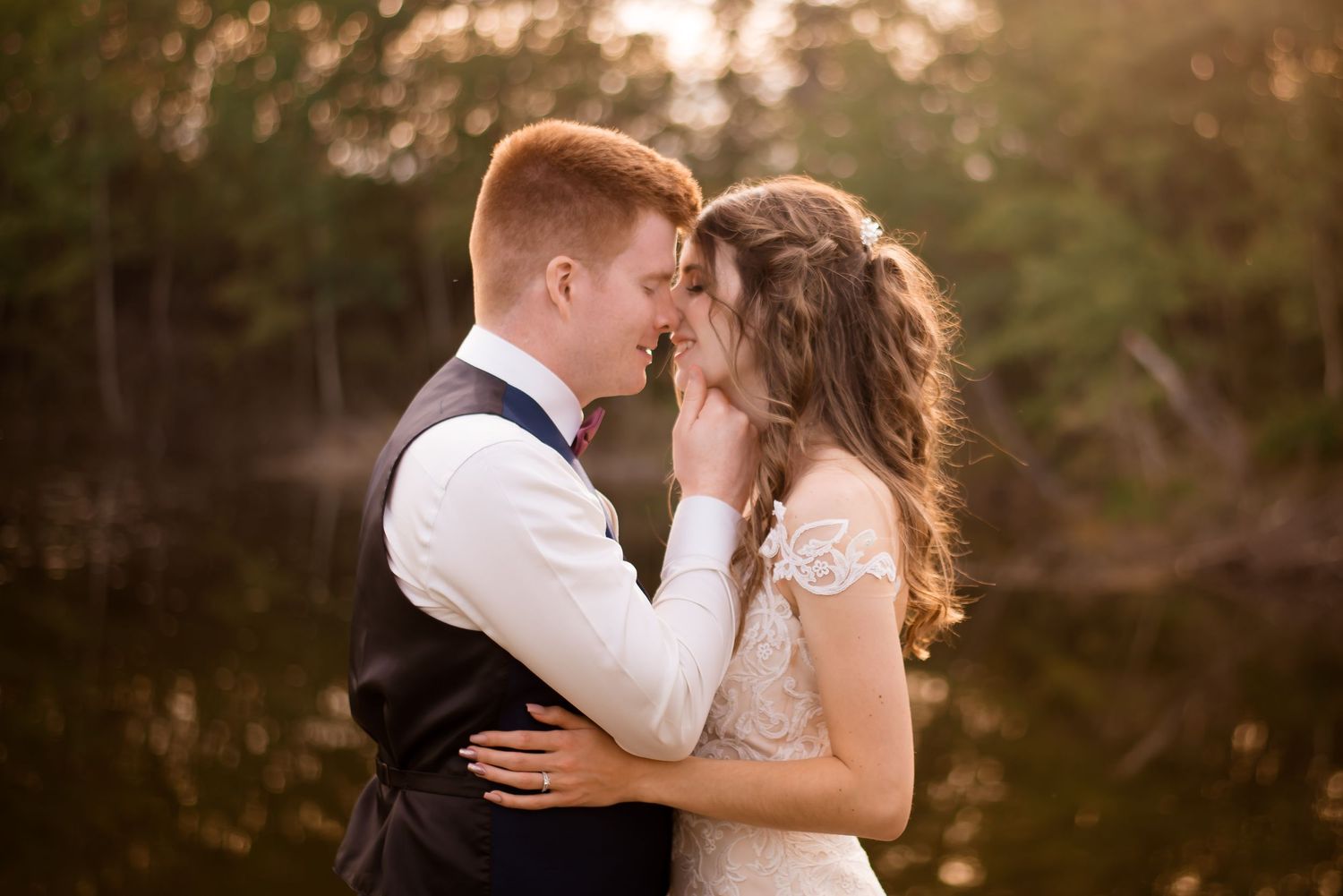 Romantic sunset wedding portrait with bride in lace gown and groom in vest embracing in golden outdoor light.