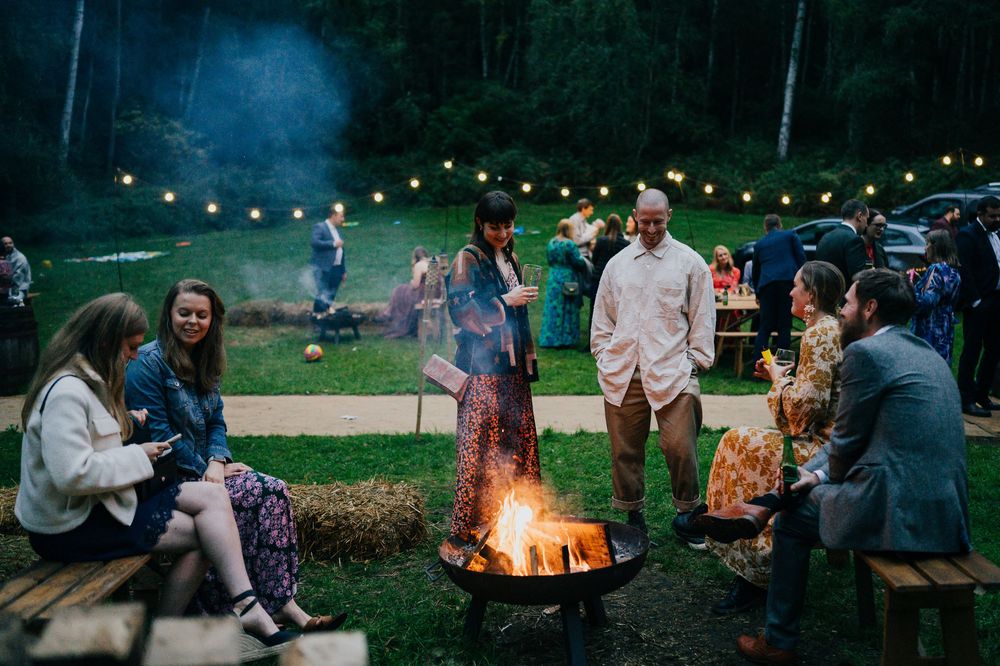 Friends gather around a cozy bonfire in an evening wedding reception at dusk enjoying drinks and conversation.