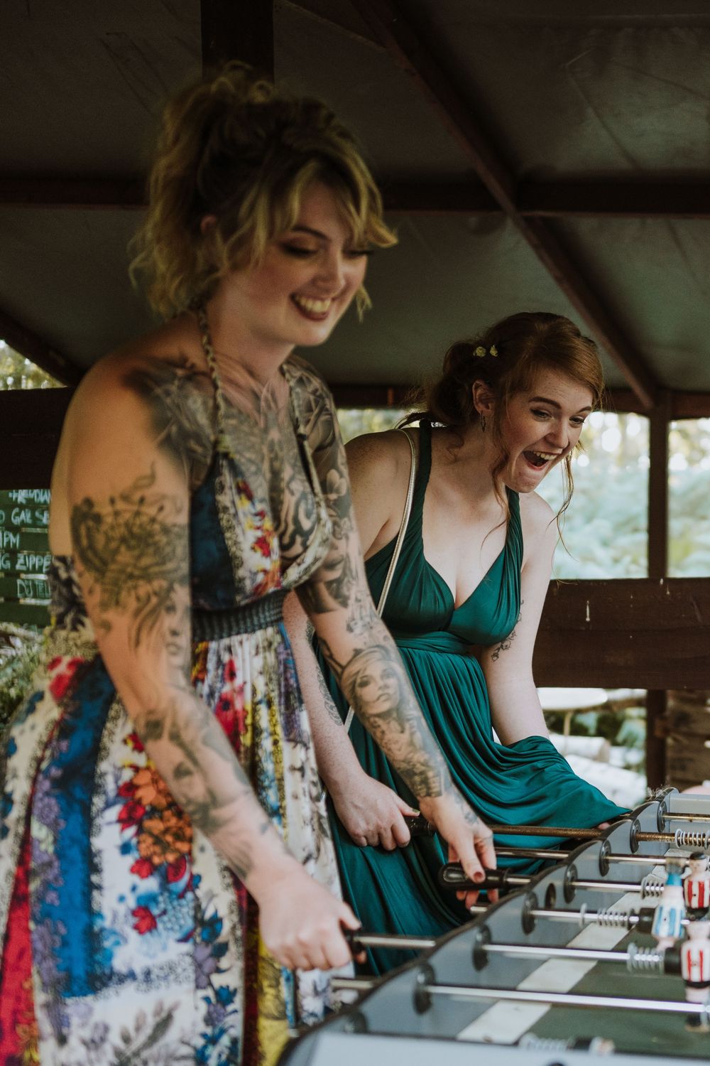 Two people in colorful dresses share a laugh while playing foosball at an outdoor venue.