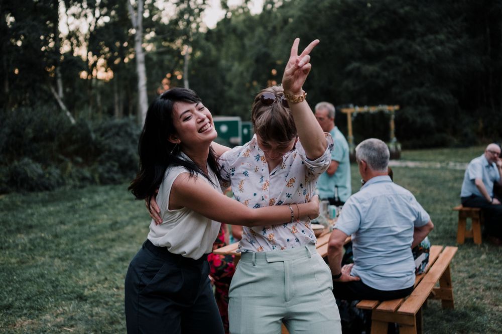 Two people dance joyfully at an outdoor gathering with picnic tables and trees in the background during sunset.