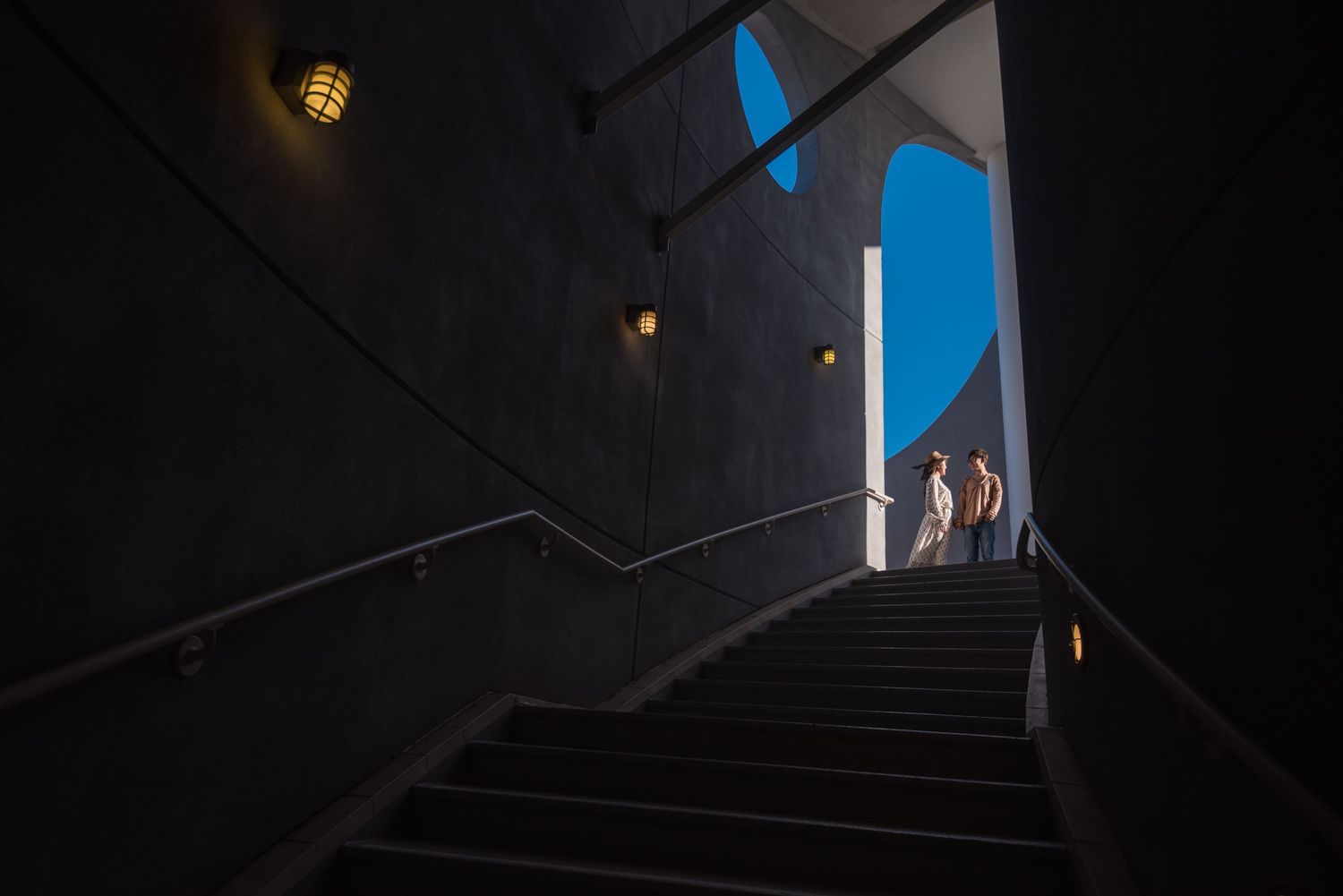 Dark stairway leading up to bright blue opening with silhouettes of people visible.