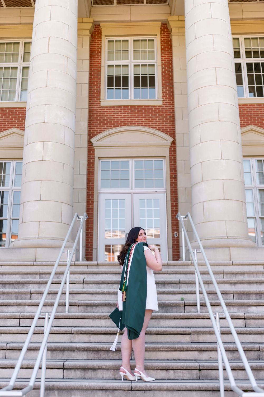 Graduate in green regalia poses on brick university building steps wearing white dress and heels holding cap.