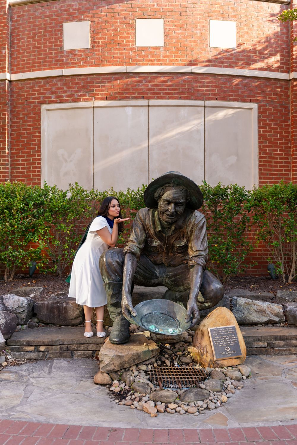 Woman in white dress posing next to a bronze gold prospector statue outside a brick building.