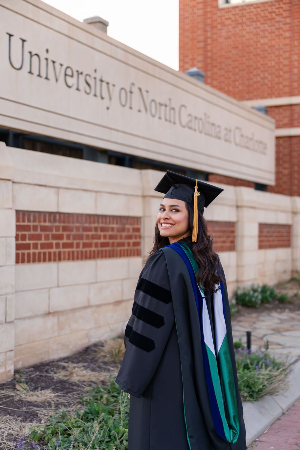 Woman in doctoral graduation gown and cap smiling in front of University of North Carolina at Charlotte sign.