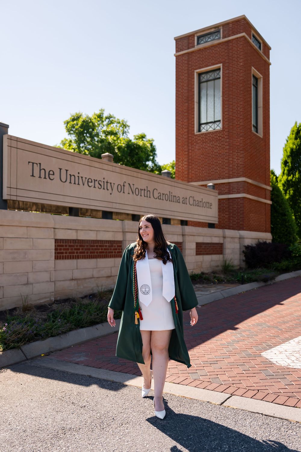 Graduate in green cap and gown poses at University of North Carolina Charlotte campus entrance sign.
