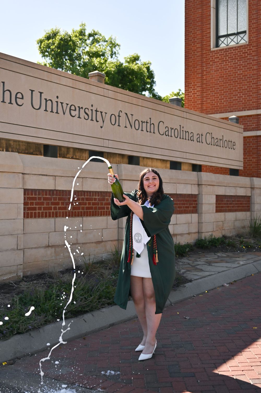 Graduate in green cap and gown celebrates with champagne in front of UNC Charlotte sign.