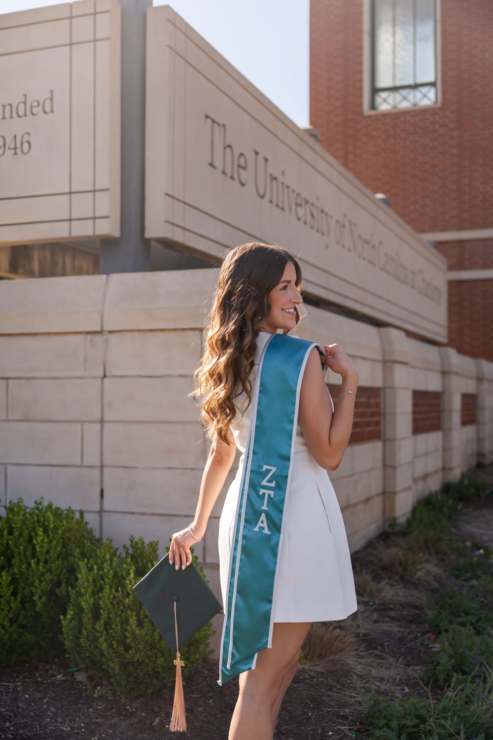 Smiling graduate in white dress wearing ZTA sash holds cap outside University of North Carolina building.