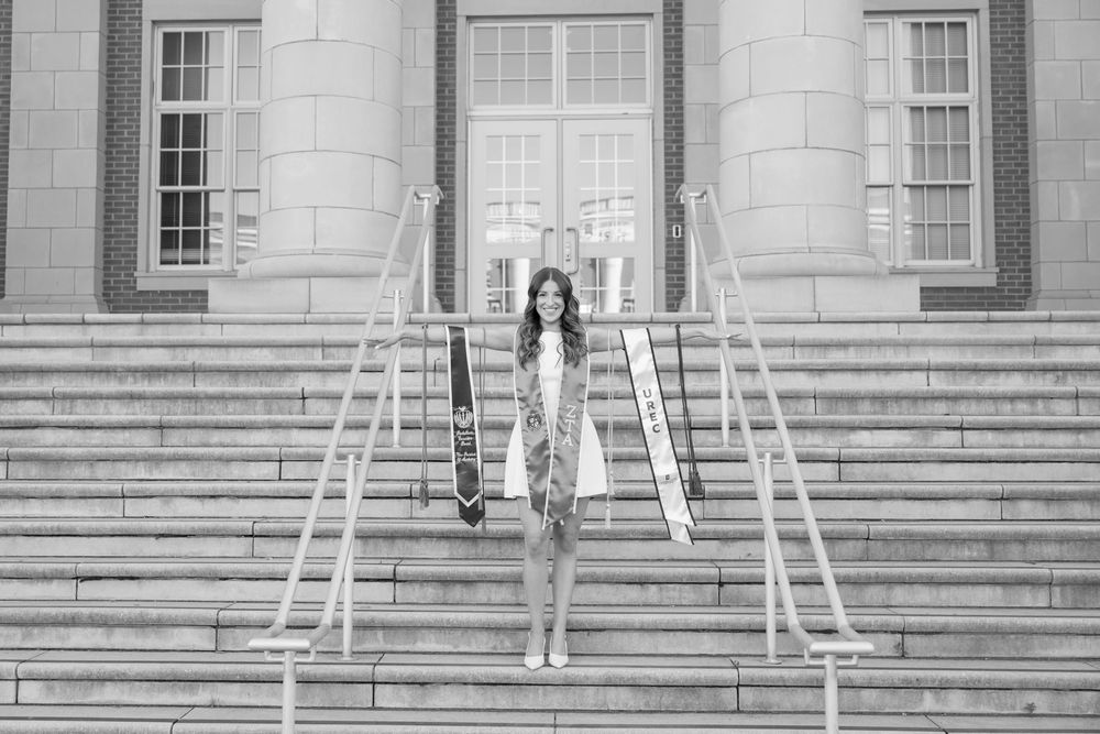 Young woman in graduation attire standing on steps of a grand building in black and white photo.