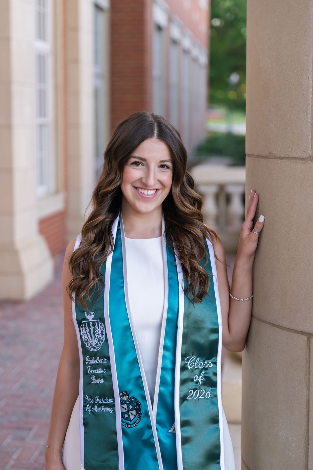Smiling college graduate wearing a teal Class of 2026 graduation stole, posing by a brick column on campus.