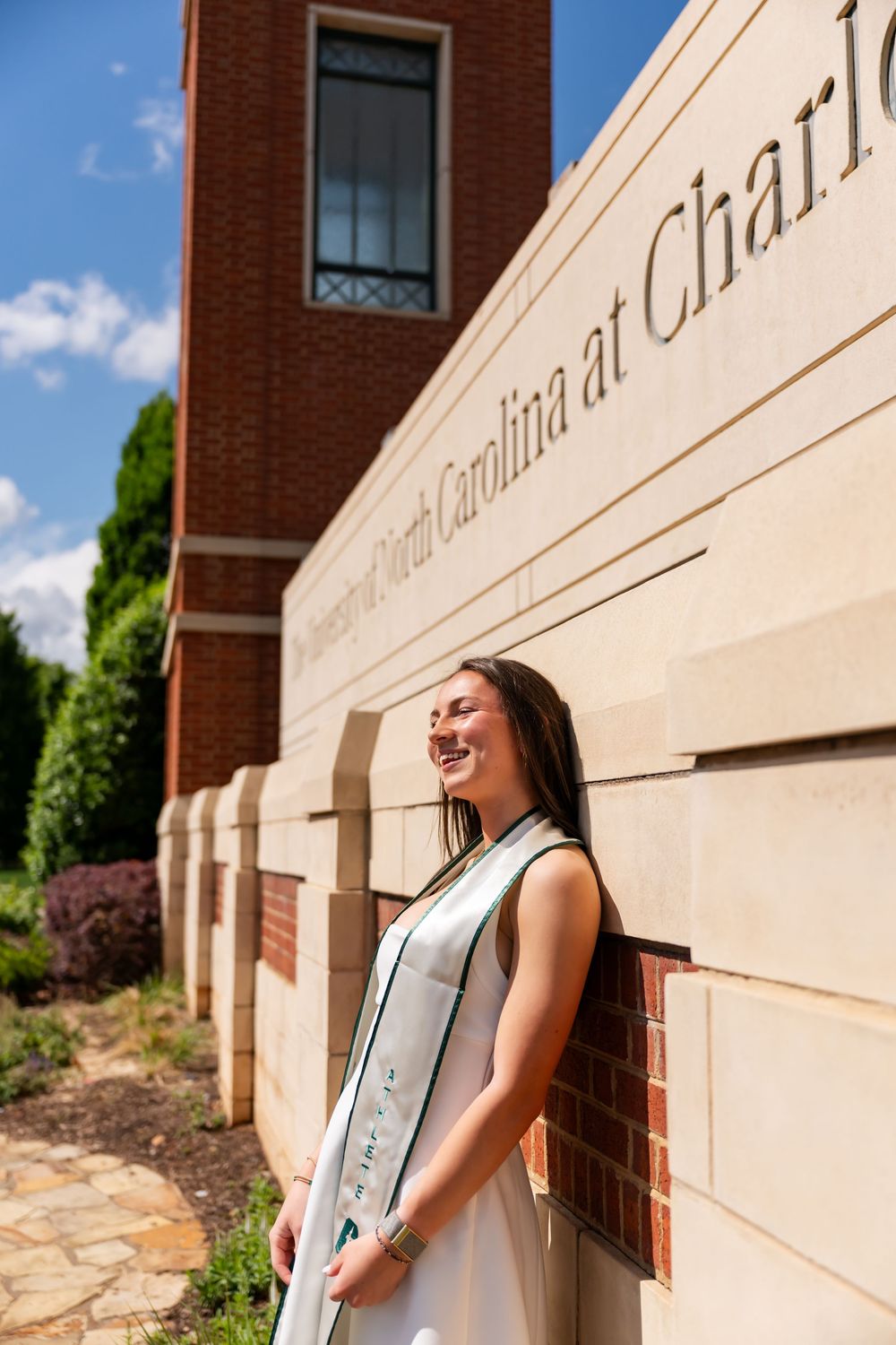Smiling graduate in white gown poses beside University of North Carolina at Charlotte campus sign on sunny day.