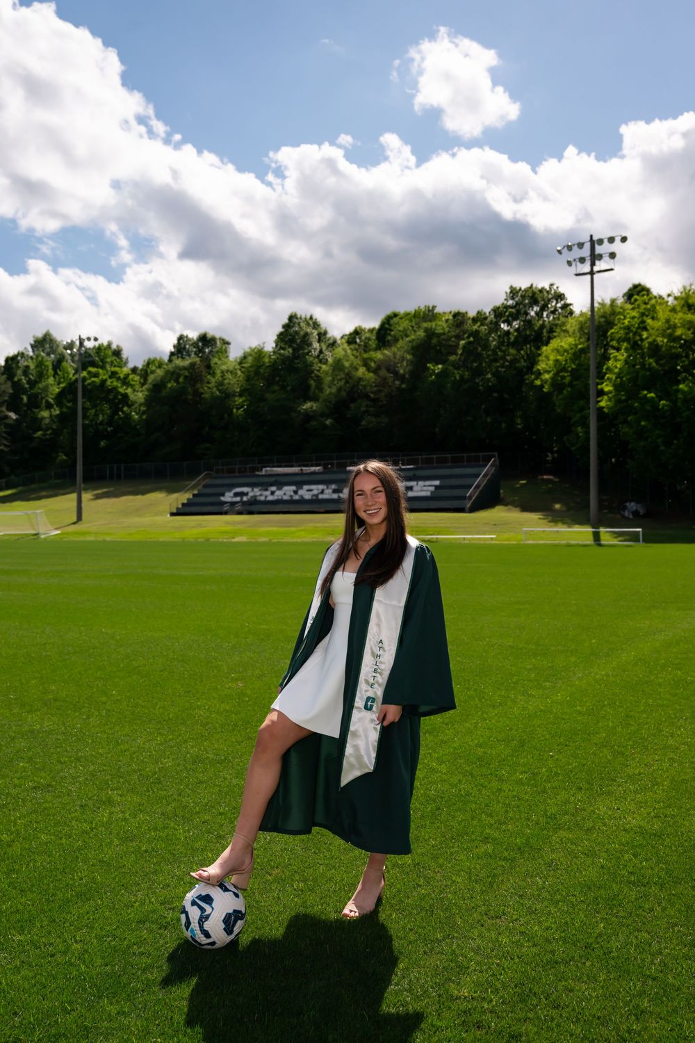 Female graduate in green cap and gown poses with soccer ball on field.