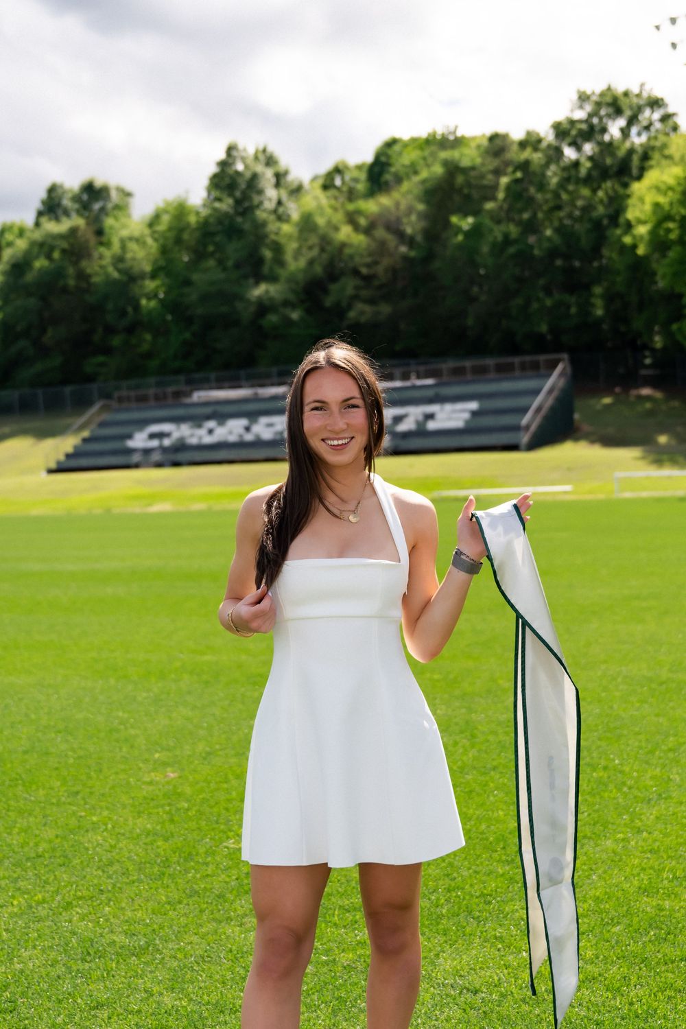 Smiling young woman in white mini dress holding a ribbon on a green sports field with bleachers behind her.