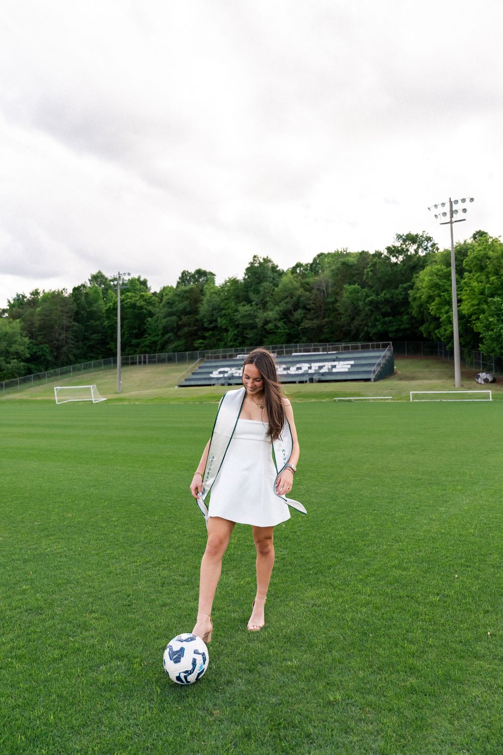 Young woman in white dress kicking soccer ball on green field at Charlotte stadium.