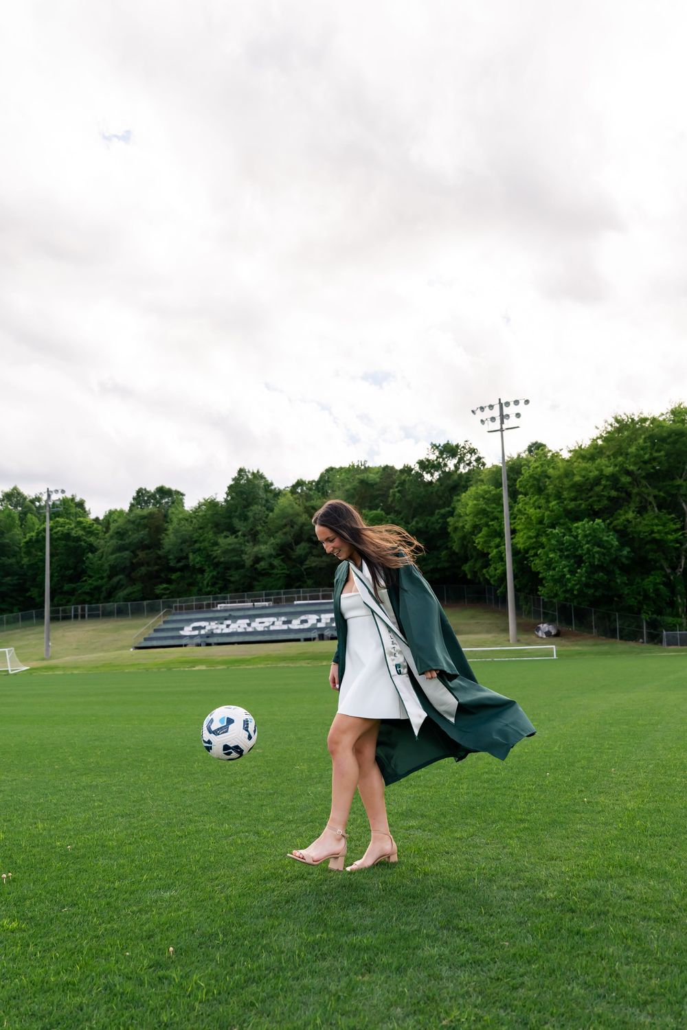 Graduate in green gown kicks soccer ball barefoot on green field.