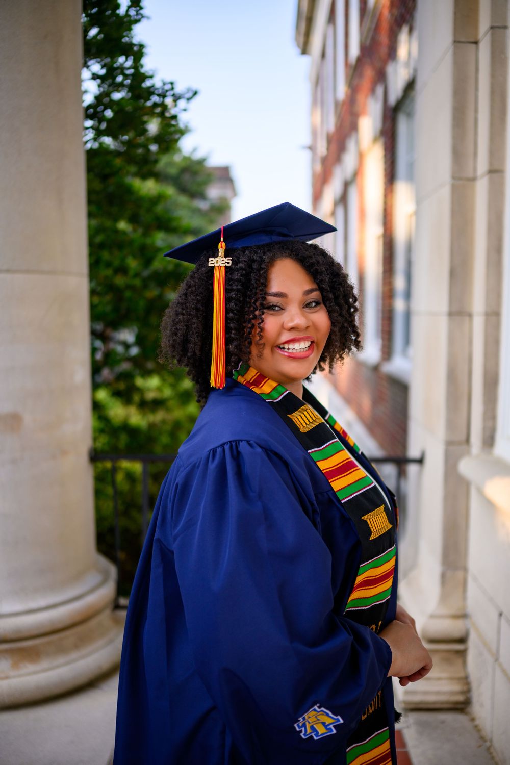 Graduate in blue cap and gown with colorful kente stole poses against architectural columns in graduation photos.