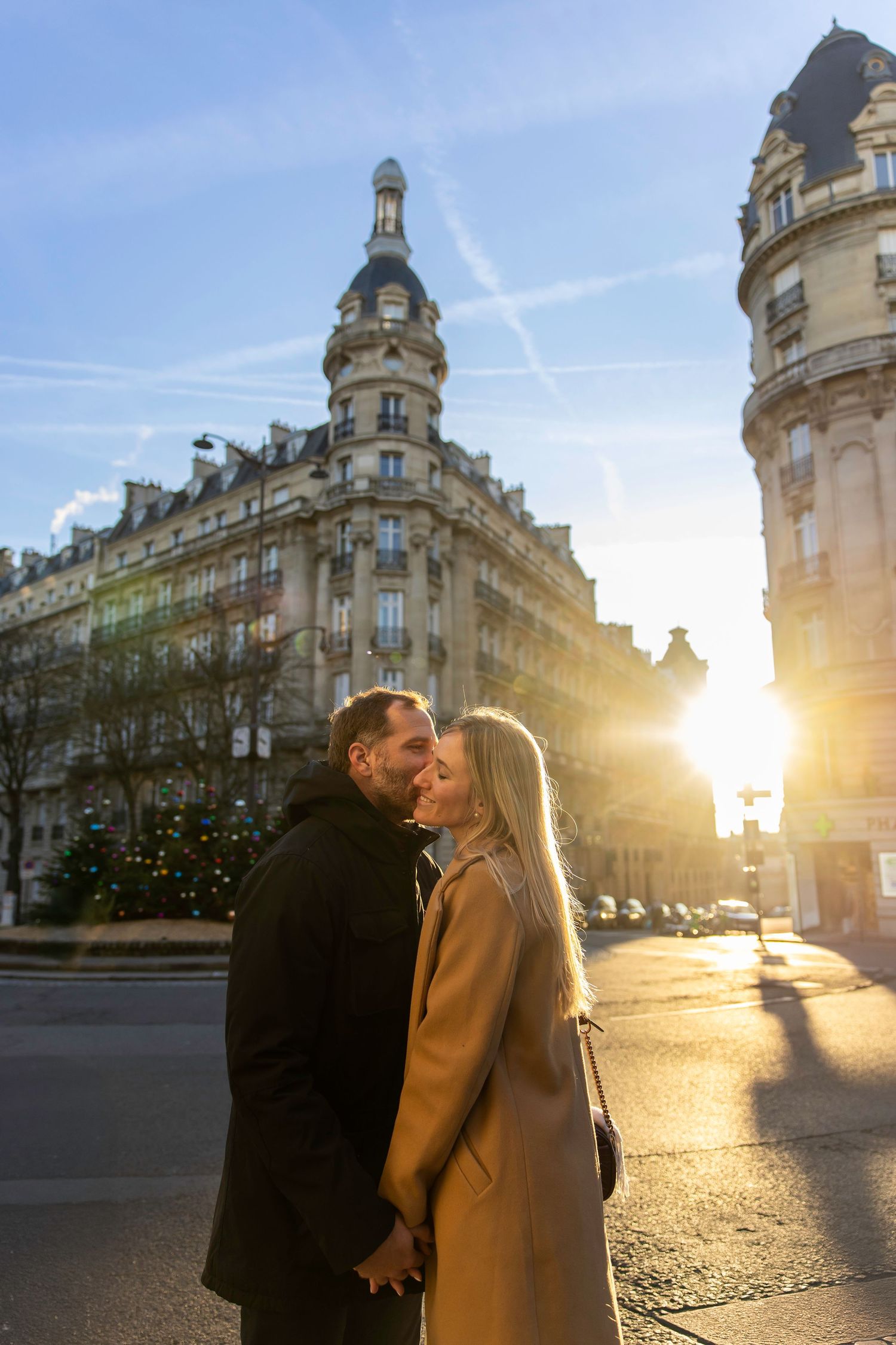 Romantic couple shares tender moment at sunset on a Paris street corner with elegant architecture and sunburst in background.