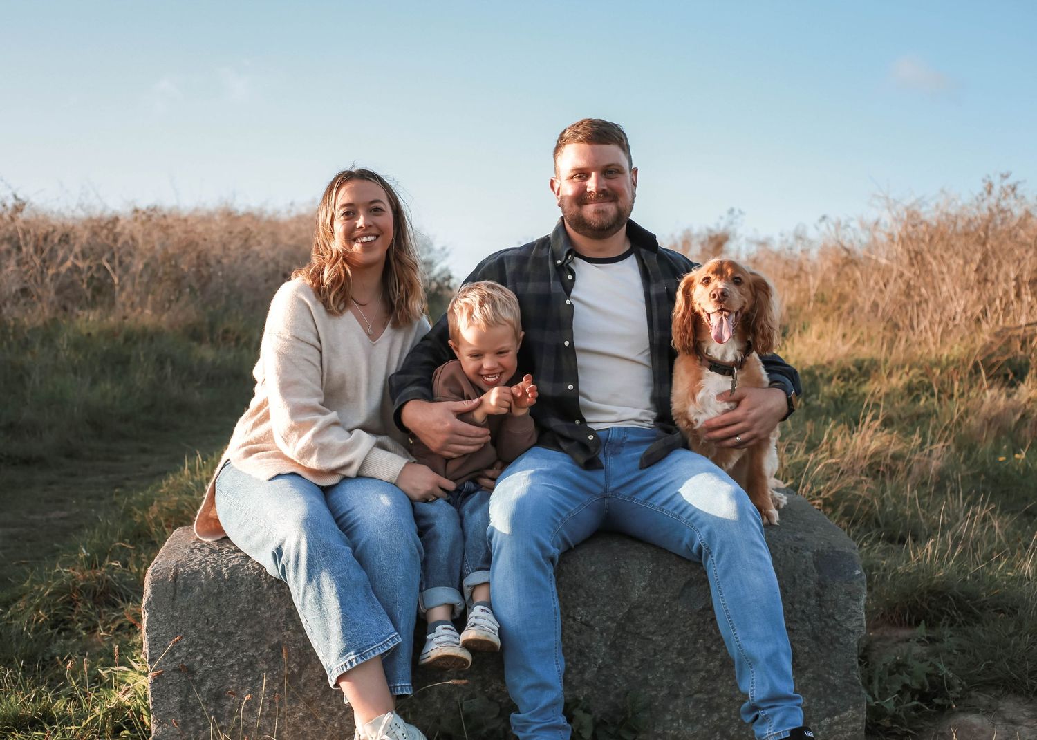 A couple sitting in a grassy field at sunset petting their cocker spaniel dog.