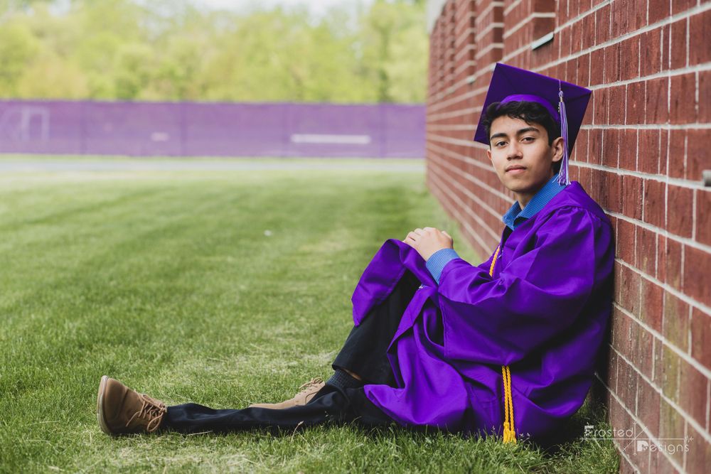 Male boy high school senior photography in purple cap and gown against red brick wall.