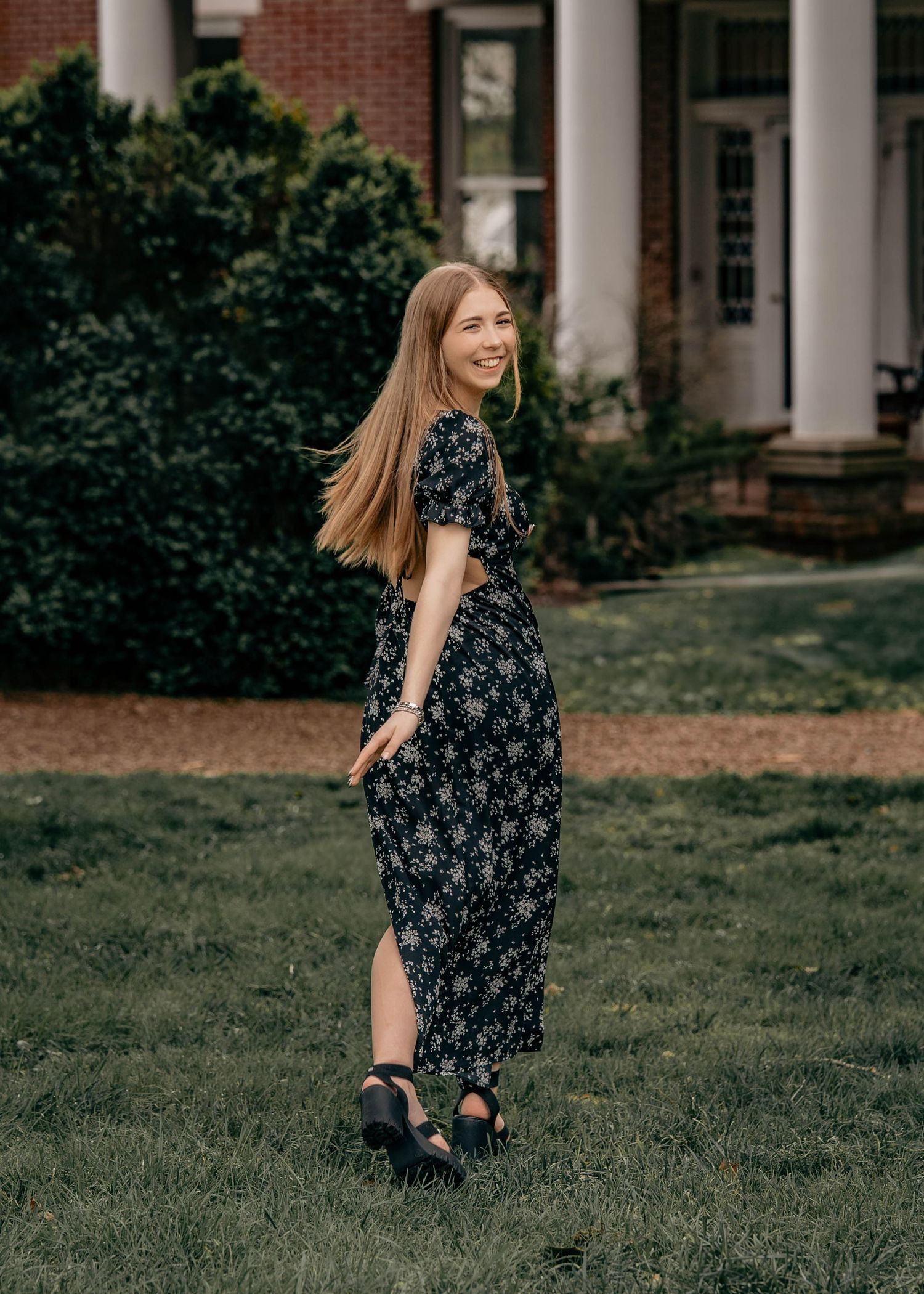 A woman in a navy floral dress stands on a lawn in front of a brick building with white columns.