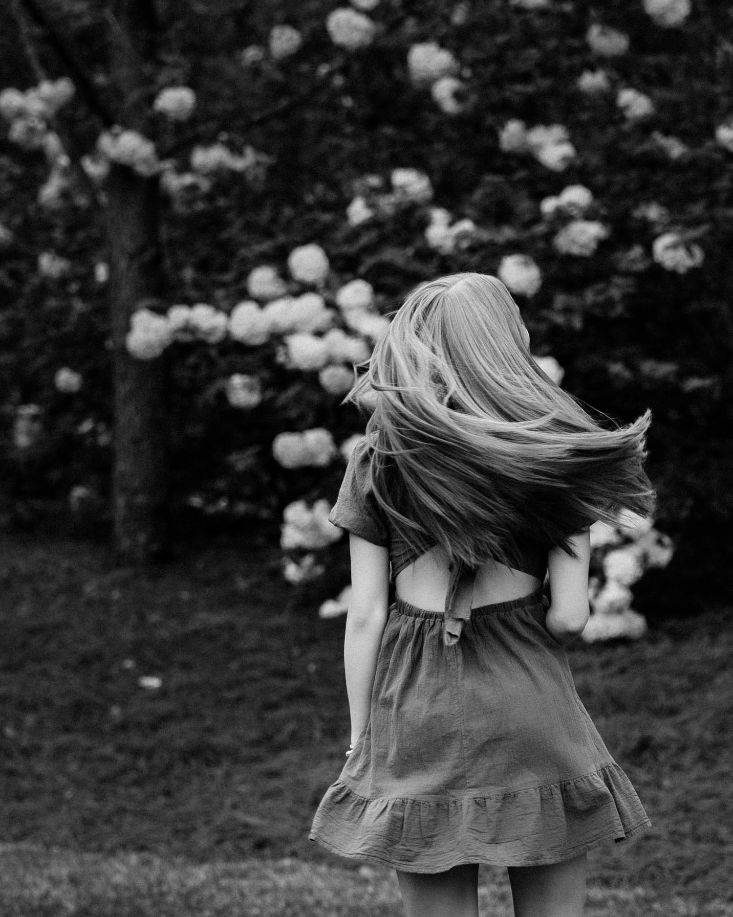 Black and white photo of a twirling figure in a dress amid blooming flowers in a garden setting.