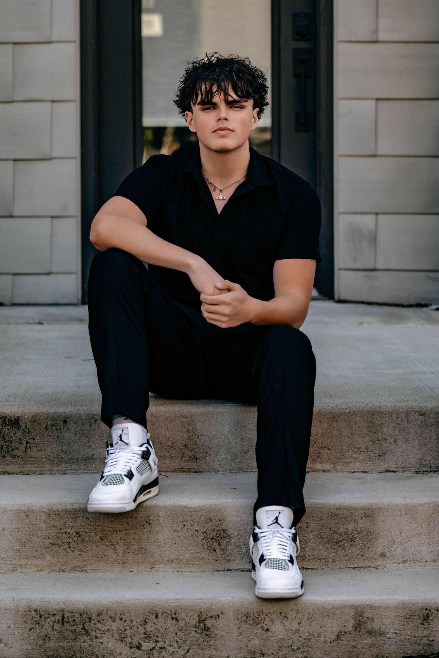 A person in all black clothing sits on concrete steps wearing white Jordan sneakers and sunglasses in an urban setting.