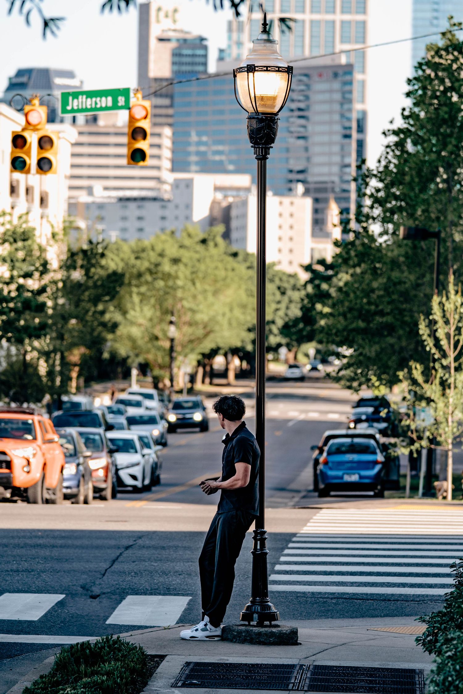 Person standing by vintage-style lamp post on urban street corner with city buildings and traffic lights in background.