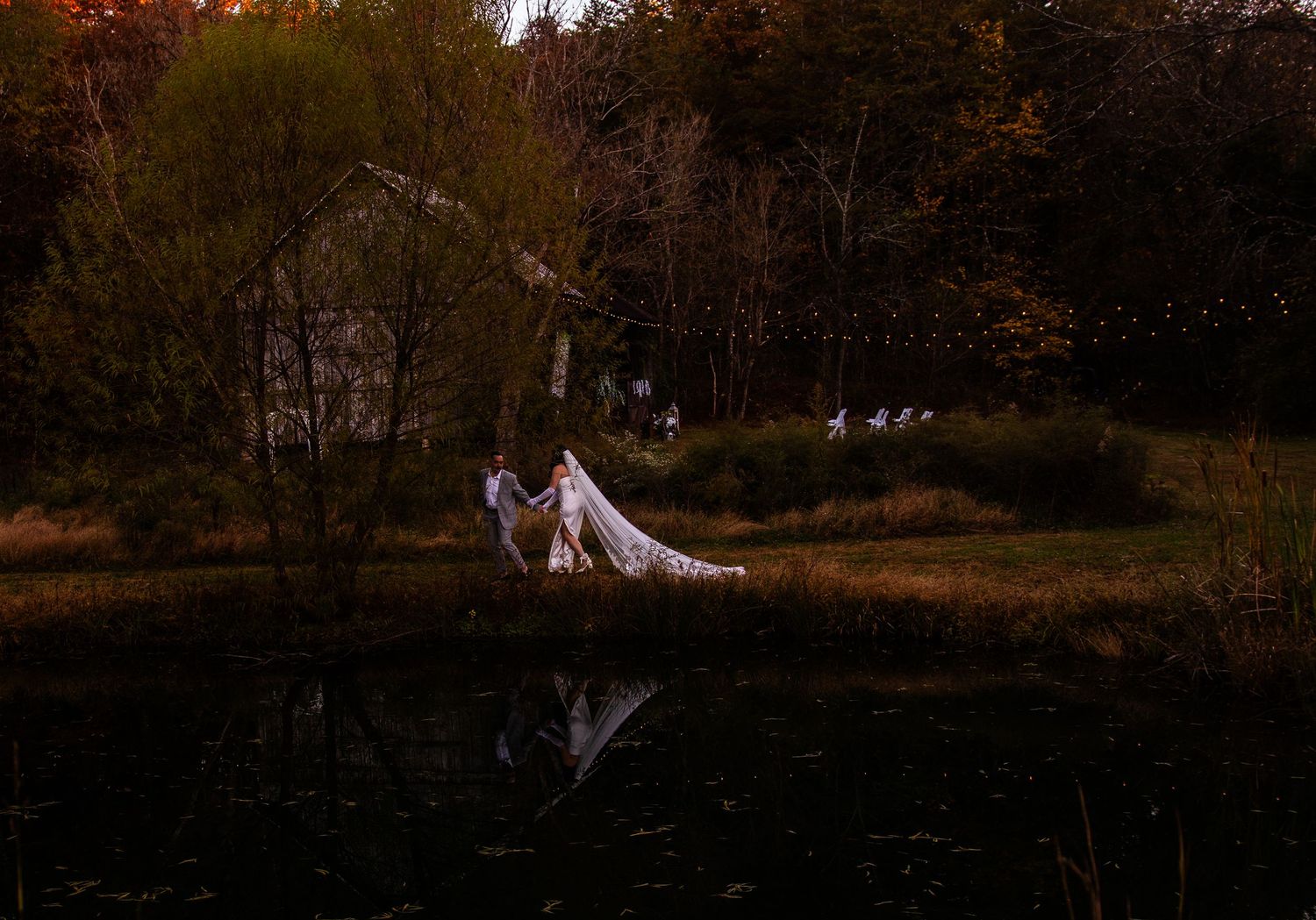 A couple stands on a wooden dock over a still pond surrounded by autumn trees.