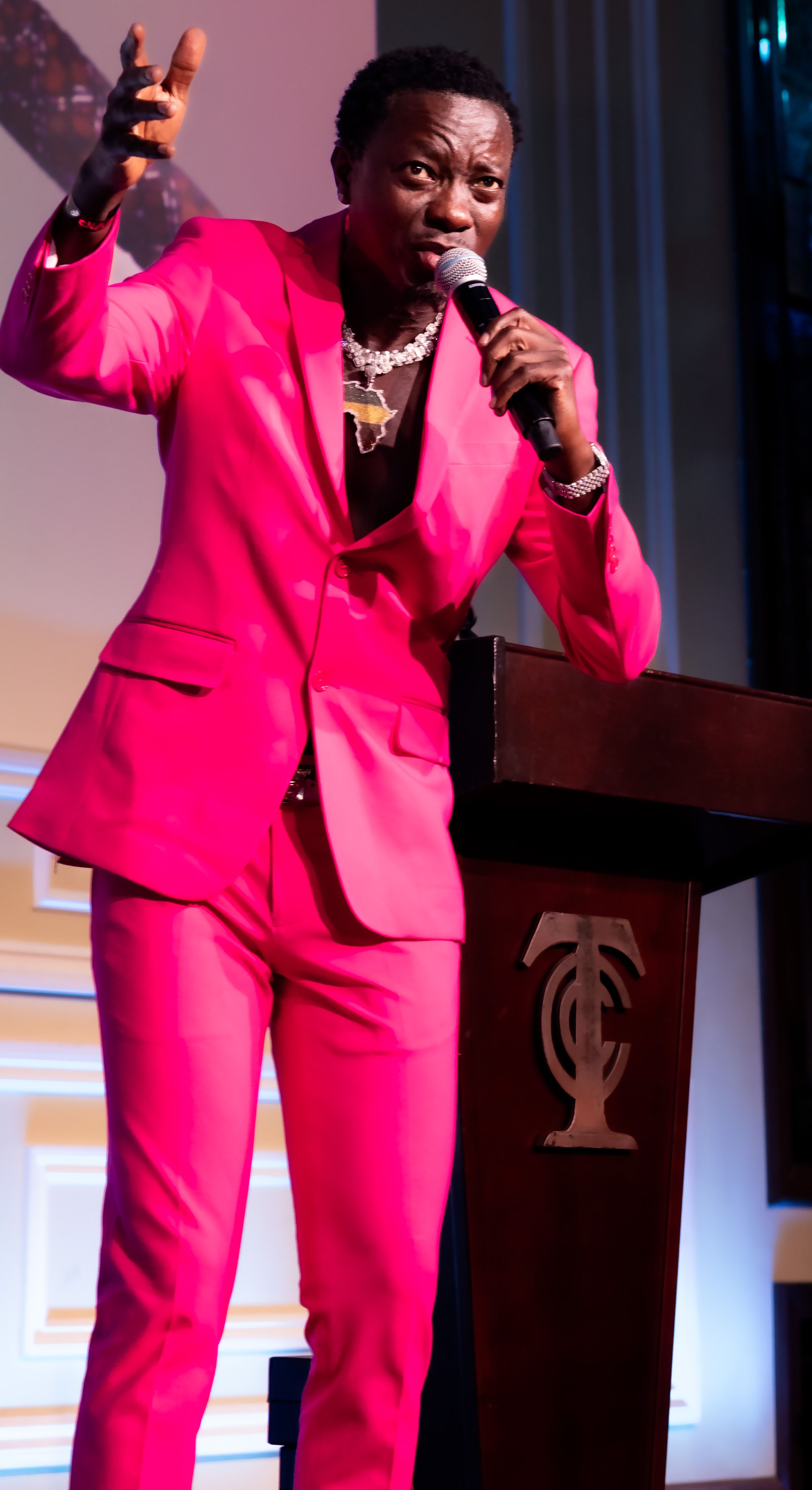 Speaker in bright pink suit addresses audience from podium at formal event.