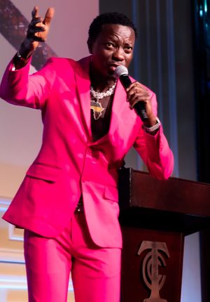 Performer in vibrant hot pink suit gestures expressively while speaking at podium during event.