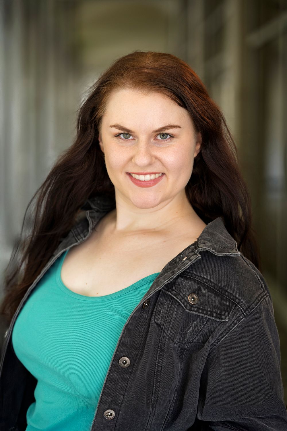 Smiling young woman with long brown hair wearing a teal top and black denim jacket poses outdoors.