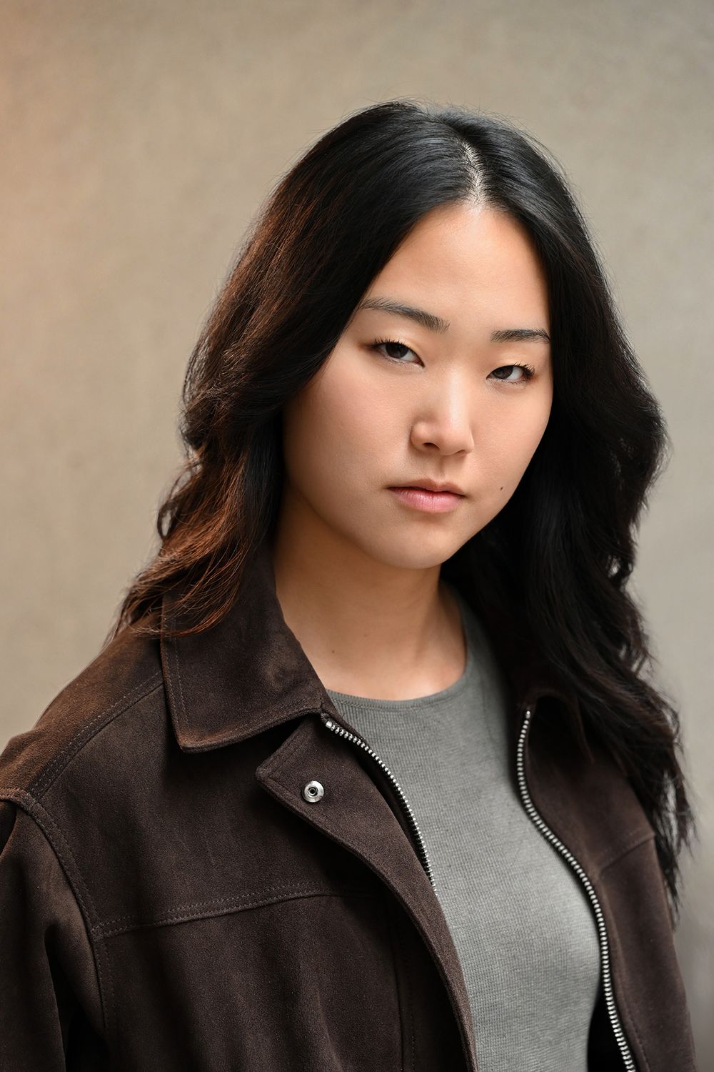 Young Asian woman with long dark hair wearing a brown jacket and gray shirt, posing for a portrait photo.