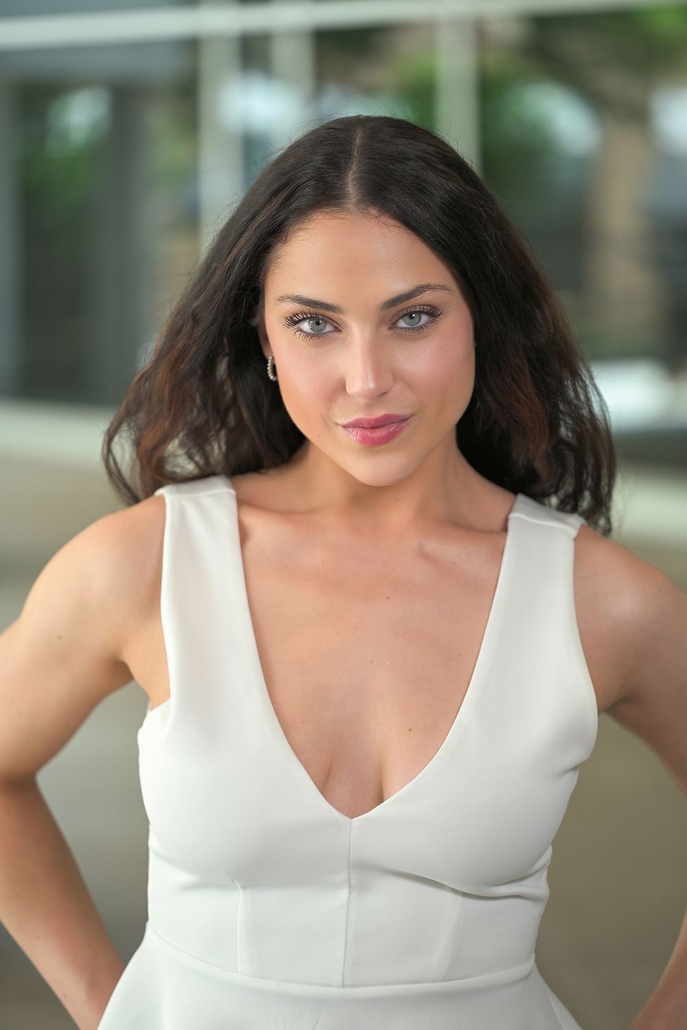 Young woman with dark hair wearing a white sleeveless dress, posing confidently with hands on hips outdoors.