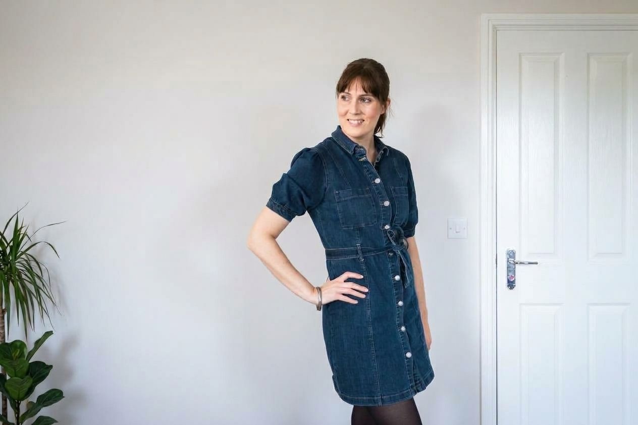 Professional photographer wearing denim dress stands confidently against white wall.