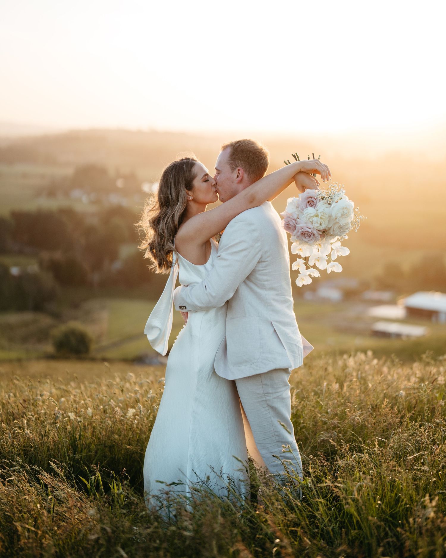 A romantic wedding couple embraces in a golden sunset field while holding a white bouquet of flowers.
