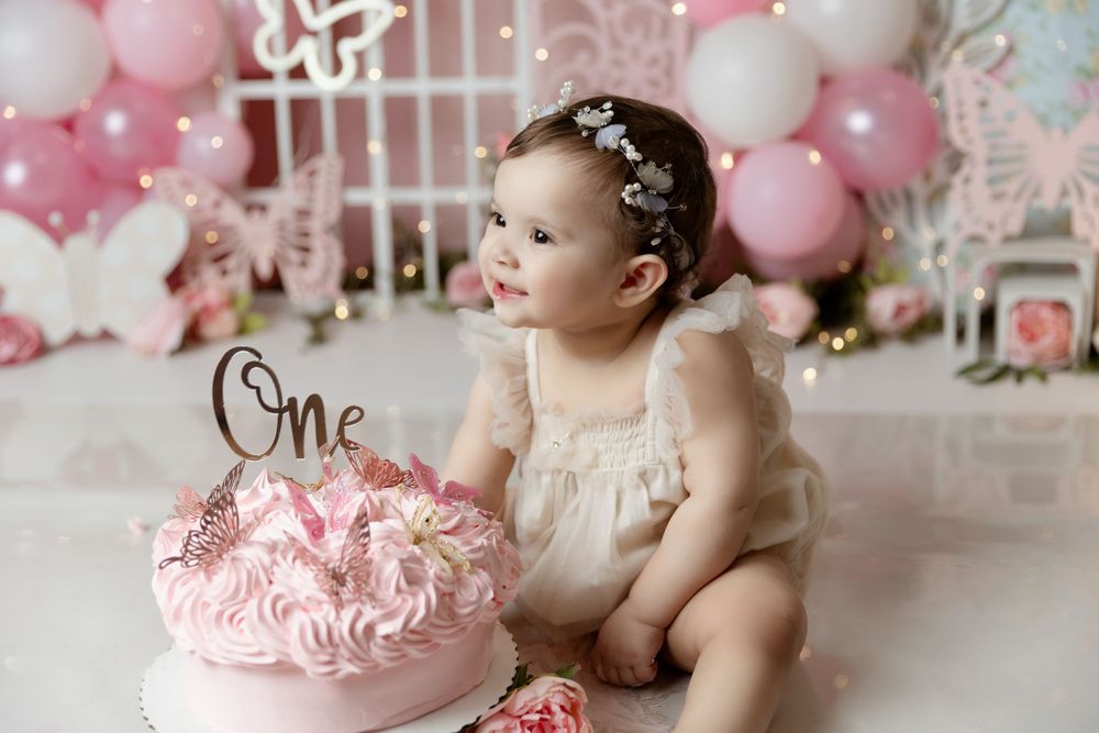 Baby in white dress near pink birthday cake with balloons and butterfly decorations.
