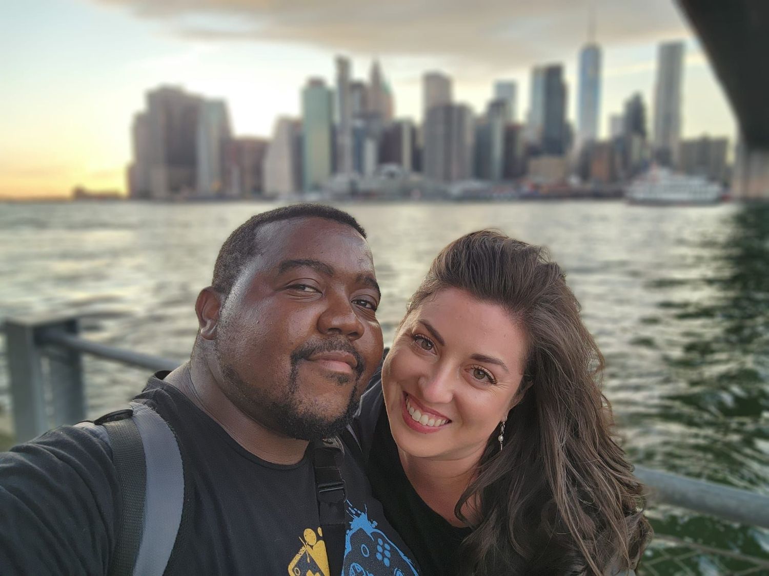 Smiling couple takes a selfie with the New York City skyline and Hudson River at sunset behind them.