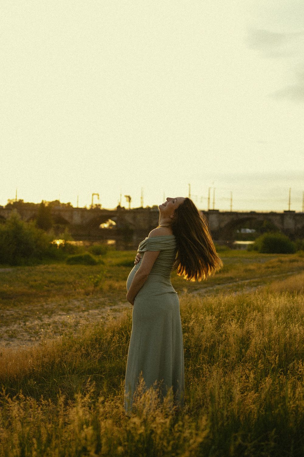 Portrait Babybauchfotoshooting an den Elbwiesen in Dresden bei Sonnenuntergang - Frau hält sich schwangeren Bauch und schüttelt Haare