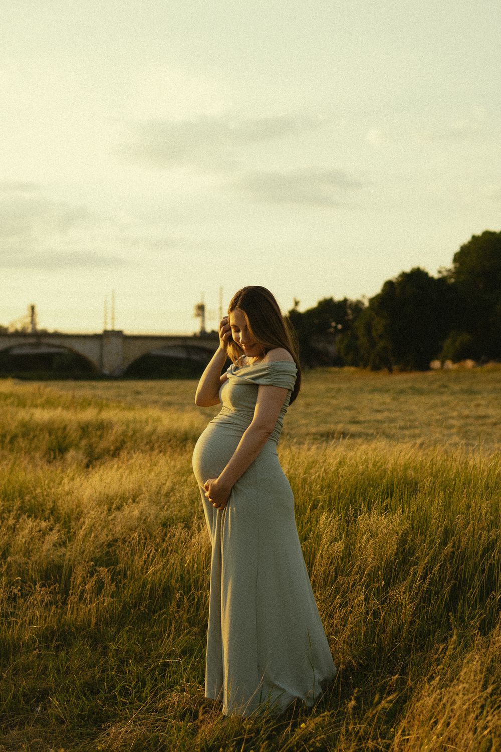 Frau hält sich schwangeren Bauch und streicht sich durch die Haare im warmen Abendrot  - Babybauchfotoshooting  Elbwiesen Dresden