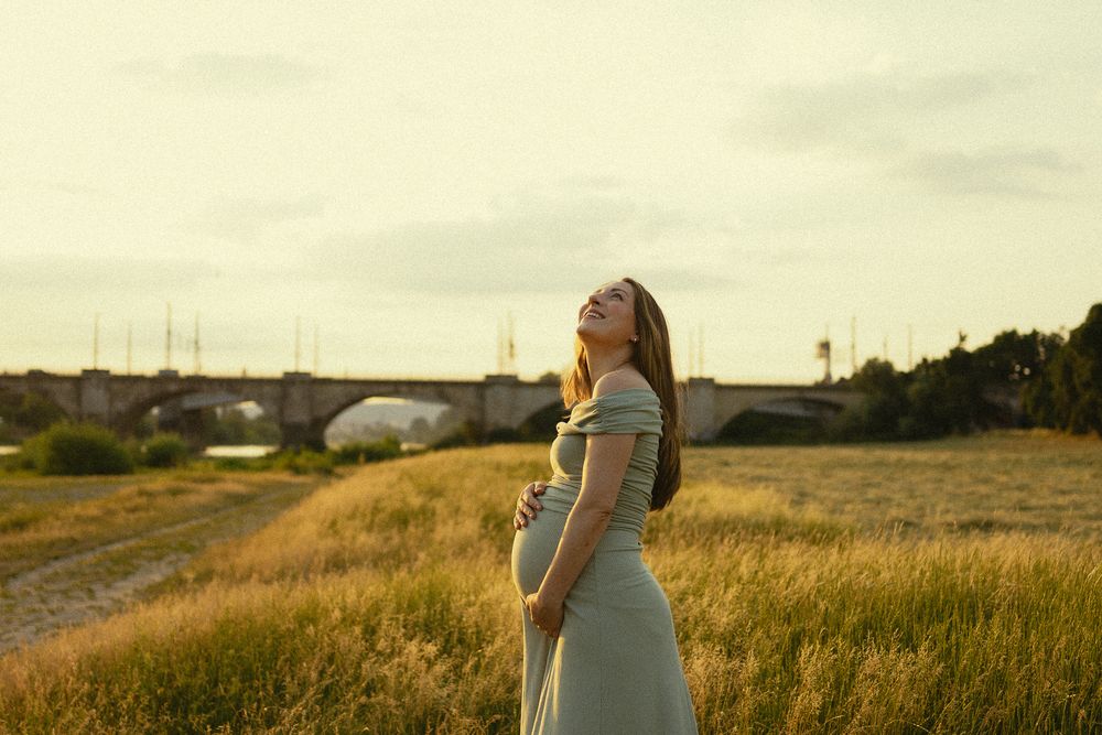 Babybauchfotoshooting Sonnenuntergang Elbwiesen Dresden - Schwangere Frau hält ihren Bauch fest und neigt Kopf in den Himmel und lacht