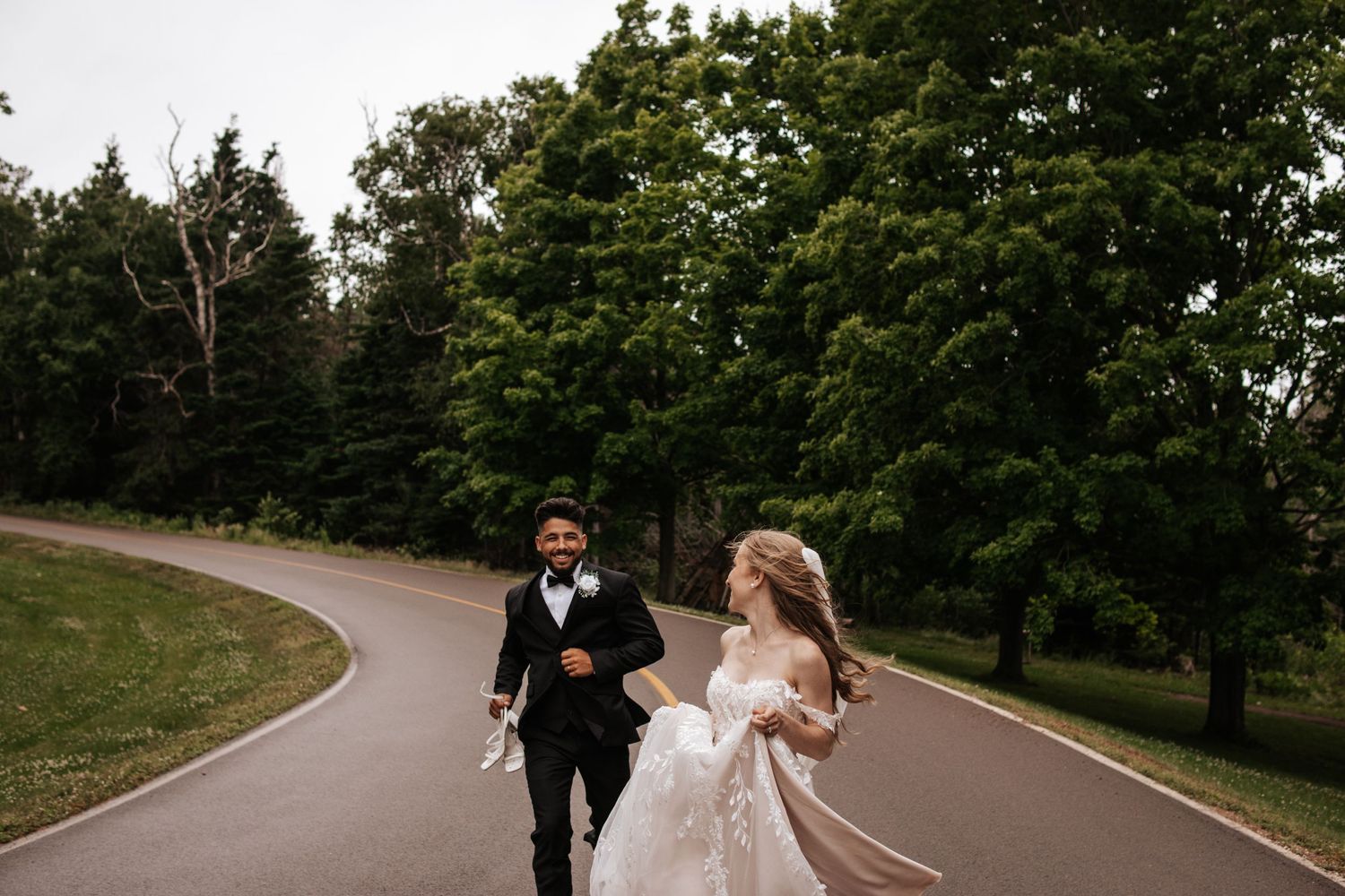 A couple in wedding attire runs joyfully down a winding path surrounded by lush green trees.