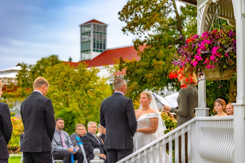 A wedding ceremony takes place on a white gazebo deck surrounded by lush greenery and bright pink flowers.