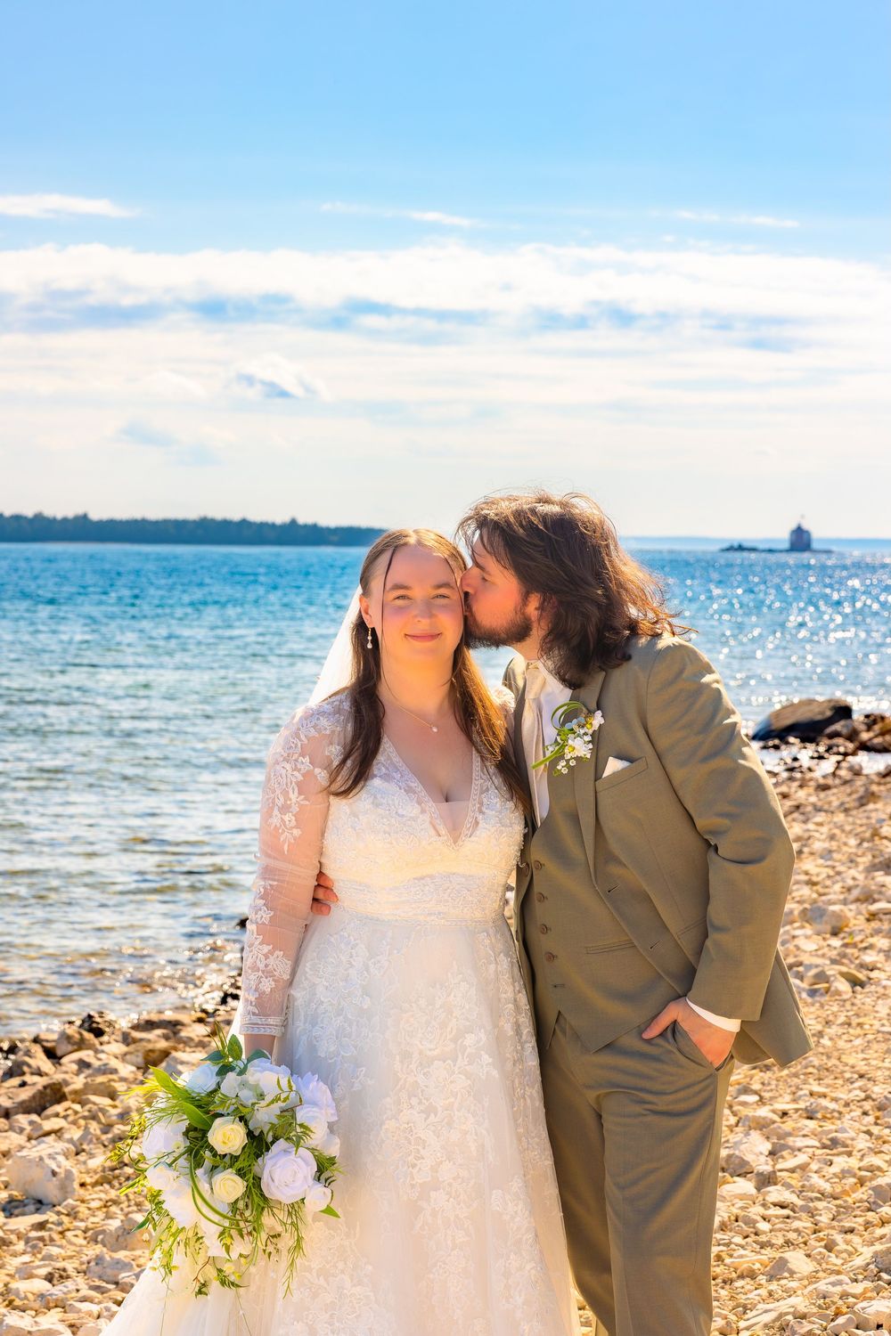 A couple shares a romantic moment on a sunny beach with sparkling blue water and rocky shoreline in the background.